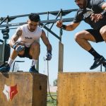 Two men demonstrating intense box jump exercises at an outdoor fitness event.